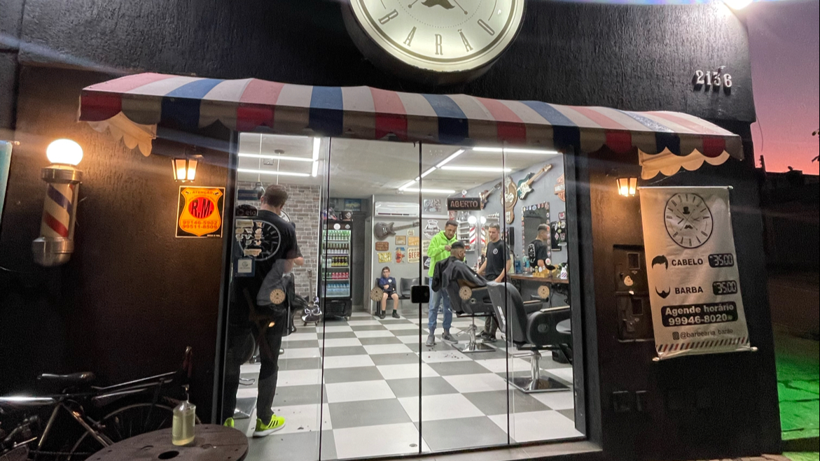 Fachada iluminada da Barbearia Barão à noite, com toldo listrado e clientes sendo atendidos, no Bairro Alto em Curitiba